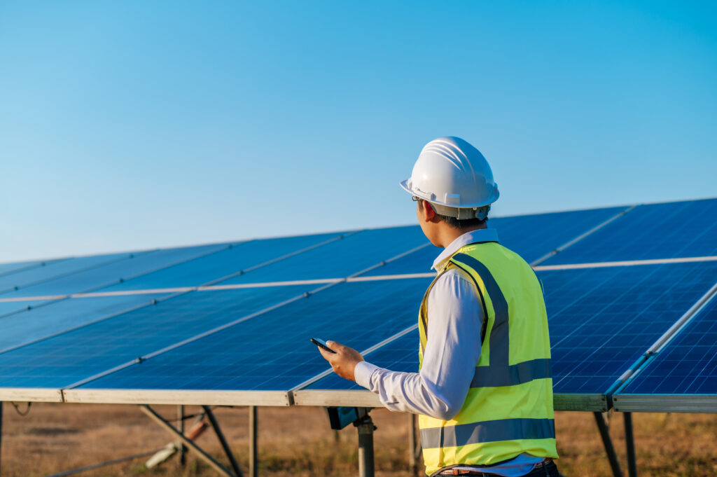 Young Asian technician man standing and talking on smartphone between long rows of photovoltaic solar panels, copy space